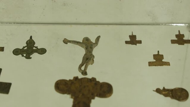 Ancient details of pectoral crosses in museum, close-up. Old crucifix figurine and metal symbols of faith lie on illuminated transparent shelf on archaeological exhibition.