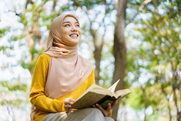 Happy cheerful Asian beautiful Muslim woman reading a book - Koran under the tree. Female muslim student studying an Islam Koran.