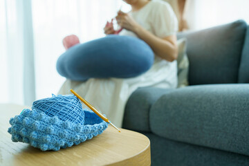 Fototapeta premium Happy cheerful Asian woman doing a crochet in living room in free time. Skillful Asian craftswoman knitting a handcraft crochet sweater and hat in cozy living room.
