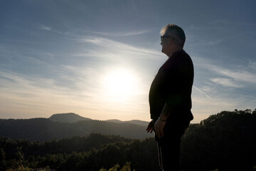 man dressed in black with sunglasses, short gray hair with toupee on top of a rock looking at a large landscape of mountains and forests, thoughtful and pondering