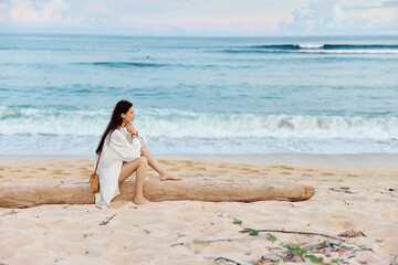 A woman with long wet hair after swimming sits on the sand with her back to the camera and looks at the sunset by the ocean on the island of Bali