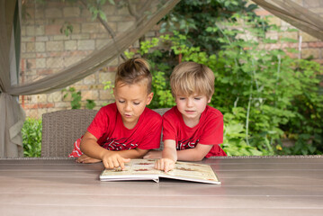 5-year-old boys in red t-shirts look with interest at pictures in open book in front of them....