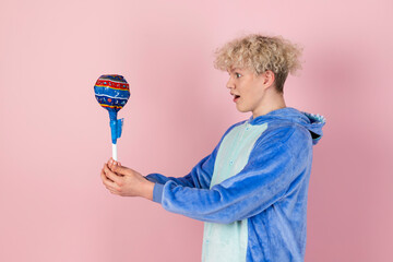 A curly blond guy in a cute and funny blue pajamas pretends to be a kid, holding a big lollipop. Isolated on pink background.