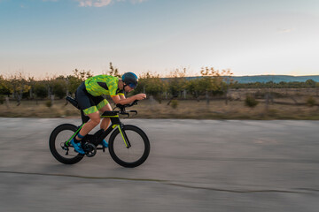 Obraz premium Triathlete riding his bicycle during sunset, preparing for a marathon. The warm colors of the sky provide a beautiful backdrop for his determined and focused effort.