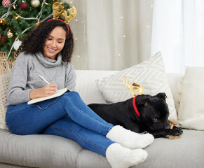 Making a list of some last minute Christmas shopping. a woman writing in her notebook while sitting at home with her dog during Christmas time.