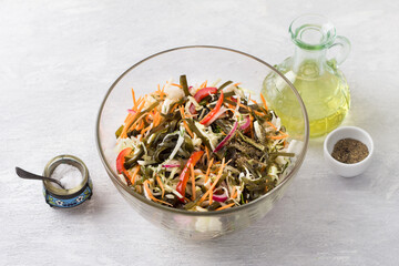 A glass bowl with a salad of white cabbage, carrots, sweet pepper, red onion, seaweed and greens surrounded by a salt shaker, a bottle of olive oil and a pepper shaker on a light gray background