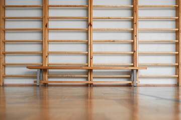 Wooden gymnastics bench stands on floor of empty gym with swedish ladder background. Sport activity exercises and physical education concept.