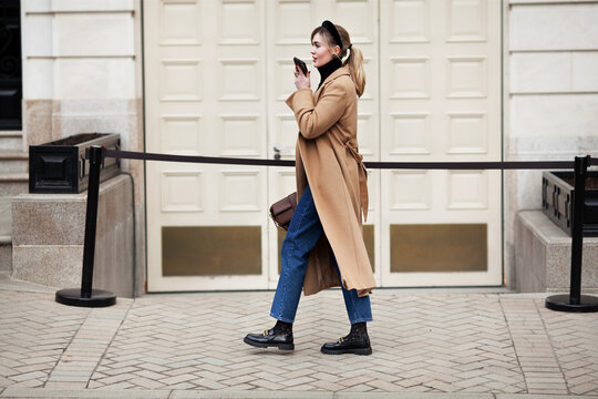 Stylish real woman walking street, using smartphone, record audio voice message or talk on loudspeaker phone. Authentic Full length Female model in motion