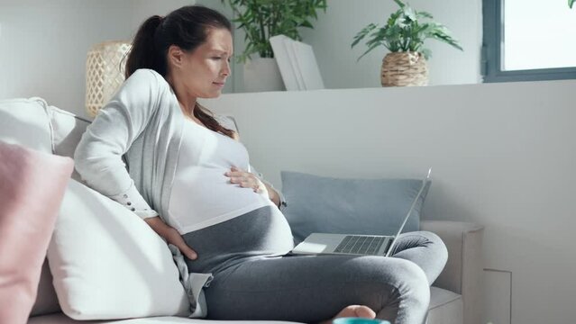 Video of worried pregnant woman with a stomachache working with laptop sitting on sofa in the living room at home