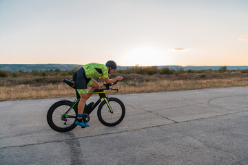 Fototapeta premium Triathlete riding his bicycle during sunset, preparing for a marathon. The warm colors of the sky provide a beautiful backdrop for his determined and focused effort.
