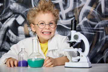 Enthusiastic cheerful boy poses table with laboratory equipment against background chalk board, looking into camera and laboratory gown. Joyful child is engaged in science studies fluids and malecules