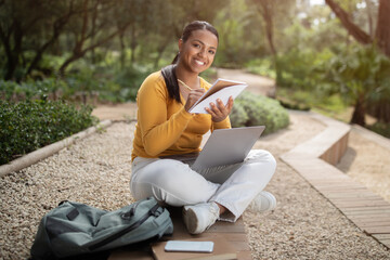 E-learning. Happy brazilian student lady preparing lesson, using laptop and taking notes, sitting outside in park