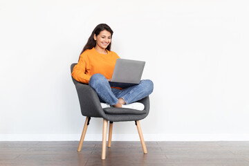 Happy pretty indian woman sitting in armchair, using laptop