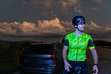 A triathlete resting on the road after a tough bike ride in the dark night, leaning on his bike in...
