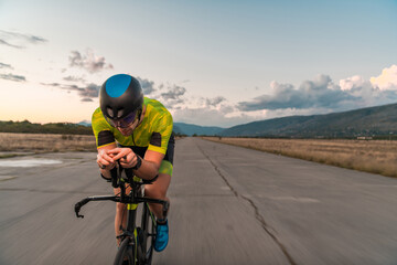 Obraz premium Triathlete riding his bicycle during sunset, preparing for a marathon. The warm colors of the sky provide a beautiful backdrop for his determined and focused effort.