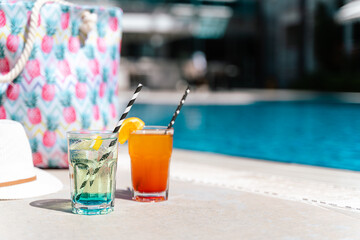 Tropical sparkling lemonade cocktails by the pool with pink beach bag and white hat in the background. Picture of glasses with orange and mint lemon fruit cocktails. Hello summer holiday vacation.