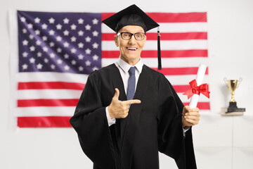 Senior man in a graduation gown holding a diploma and pointing in front of USA flag