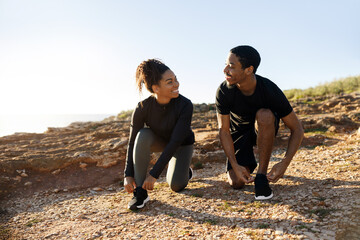 Smiling millennial african american lady and man in sportswear tying shoelaces on shoes on ocean beach - Powered by Adobe