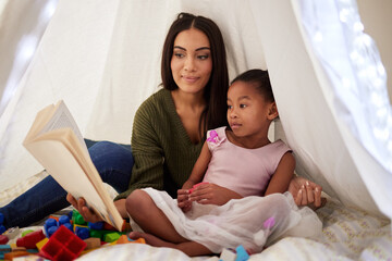 Spark your childs imagination through stories. a mother reading a book to her little daughter under a blanket fort at home.