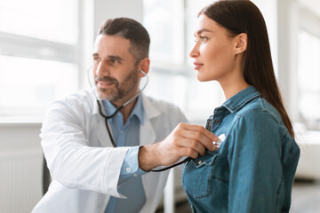 Caring male doctor using phonendoscope, examining young female patient heart rate at consultation...