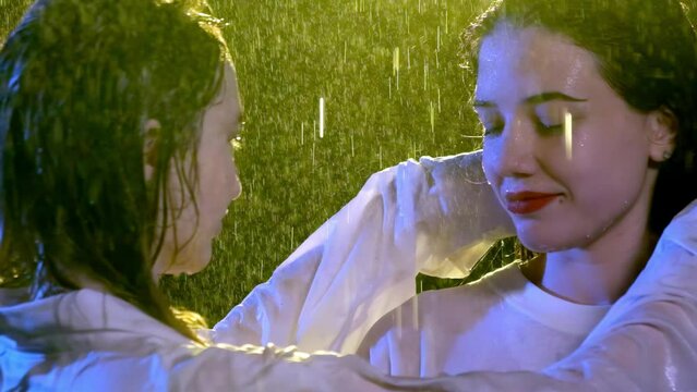Young woman adjusts wet hair of girl friend with red lipstick. Tender females in white wet shirts stand under pouring rain and warm yellow light Generative AI