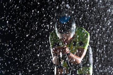 A triathlete braving the rain as he cycles through the night, preparing himself for the upcoming marathon. The blurred raindrops in the foreground and the dark, moody atmosphere in the background add