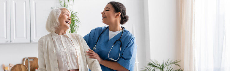 Fototapeta na wymiar happy multiracial nurse in uniform laughing with retired woman at home, banner.