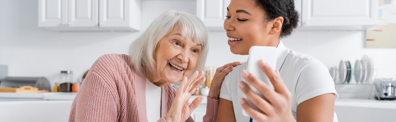 cheerful multiracial social worker holding smartphone while senior woman waving hand during video...