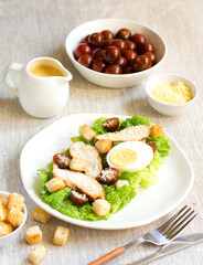 Caesar salad with croutons, fried chicken, cherry tomatoes, egg and parmesan cheese on a light plate with a fork, on a linen tablecloth in a plate of grated parmesan cheese and cherry tomatoes