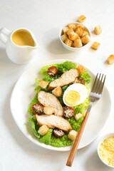 Caesar salad with croutons, fried chicken, cherry tomatoes, egg and parmesan cheese on a light plate with a fork, on a light table against the background of a bowl of croutons, grated parmesan cheese