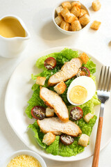 Caesar salad with croutons, fried chicken, cherry tomatoes, egg and parmesan cheese on a light plate, on a light table against the background of a bowl of croutons, grated parmesan cheese