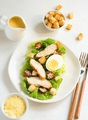 Caesar salad with croutons, fried chicken, cherry tomatoes, egg and parmesan cheese on a light plate, on a light table against the background of a bowl with croutons, grated parmesan cheese