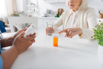 cropped view of senior woman pointing at bottle with medication near multiracial nurse on blurred foreground.