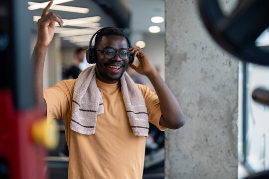 Young motivated African American sportsman listening music over headphones improving quality of workout, raising arm up, feeling happy, enjoying the rhythm in gym during workout exercise routine.
