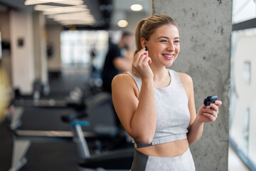 Young beautiful sportswoman with earphones. Smiling fitness woman putting on in-ear headphones, holding charging case. Happy female athlete listening to music over earphones, enjoying her time in gym.