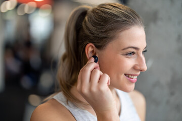 Close up photo of young beautiful sportswoman with earphones. Smiling fitness woman putting on in-ear headphones. Happy female athlete listening to music over earphones, enjoying her time in gym.