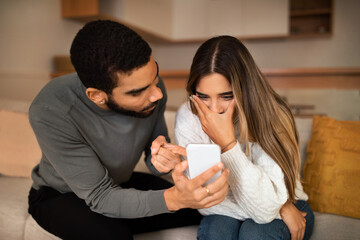 Angry millennial middle eastern husband showing smartphone to frightened caucasian wife quarreling