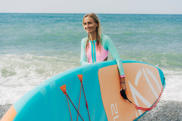 Happy smiling young woman carries sup board into the sea.