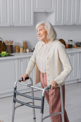 positive senior woman with grey hair walking with help of walker at home.