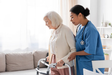 Fototapeta premium brunette multiracial nurse in uniform supporting retired woman with grey hair walking with walker at home.