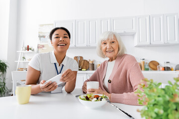 happy multiracial social worker holding smartphone during lunch with retired woman in kitchen.