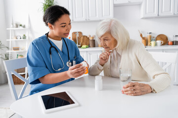 Fototapeta premium multiracial nurse showing medication to senior woman next to digital tablet on table.