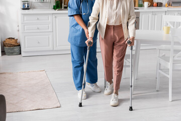 cropped view of senior woman using crutches while walking near nurse at home.