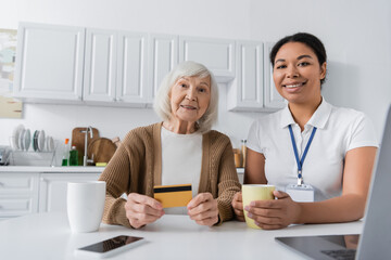 cheerful multiracial social worker holding cup near senior woman with credit card and gadgets.