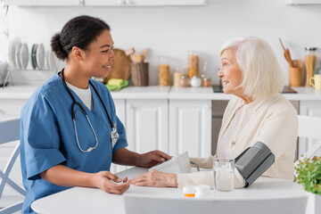 happy multiracial nurse measuring blood pressure of senior woman with grey hair next to medication on table.