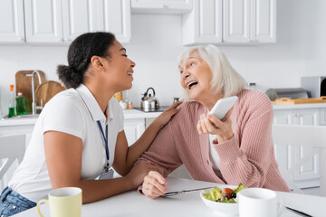 happy senior woman holding smartphone near cheerful multiracial social worker at home.