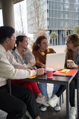 Vertical happy diverse friends study together with laptop. Classmates prepare exam drinking coffee outside campus. Group of young multiracial people using computer sitting on the terrace of cafeteria.