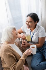 cheerful multiracial social worker chatting with senior woman while having tea in living room.