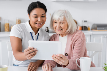 joyful multiracial social worker holding digital tablet near senior woman in kitchen.