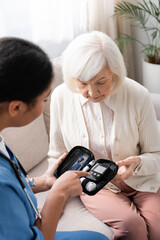 multiracial nurse in uniform pointing at diabetes kit near senior woman sitting on sofa.